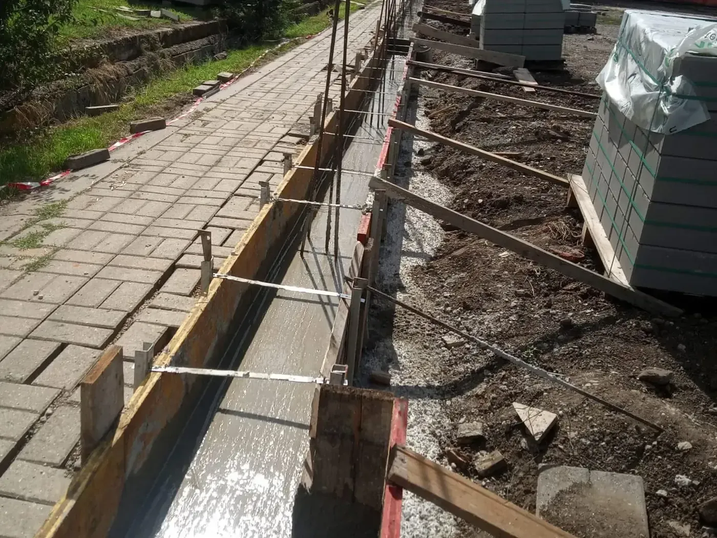 Kerítésalap zsaluzata és kitűzése járdaszegély mellett, háttérben zsalukő raklapok