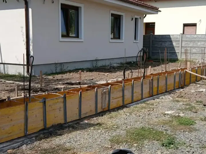 Kerítés sávalapjának fa zsaluzata vasszereléssel családi ház előtt